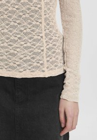 Top in pizzo a maniche lunghe di un beige chiaro con motivo floreale, indossato sopra una gonna nera. Il tessuto è delicato e testurizzato, mettendo in mostra dettagli intricati.