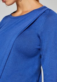 Modrý pletený svetr s kulatým výstřihem a diagonálním švem přes rameno, který vystihuje hladkou, strukturovanou tkaninu.