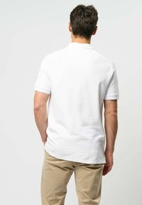 Mężczyzna stojący tyłem, ubrany w gładką białą koszulę polo z krótkim rękawem i beżowe spodnie na jednolitym jasnym tle.