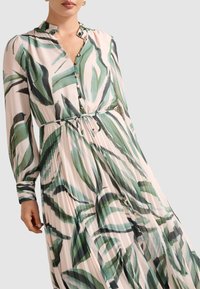 Femme portant une robe à manches longues, plissée, avec des motifs de feuilles abstraites vertes et noires et des boutons dorés sur le devant.