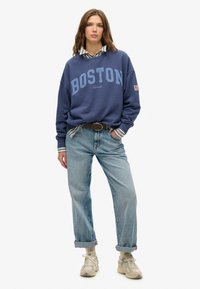 Sudadera azul marino con el logo "BOSTON", superpuesta a una camisa de cuello a rayas, combinada con unos jeans claros arremangados y zapatillas beige.