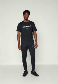 Bărbat în picioare, purtând un tricou negru Nike Spurs, pantaloni scurți negri Nike și adidași negri, pe un fundal simplu deschis.