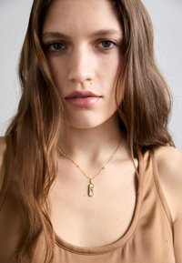 Collana con pendente d'oro con un ciondolo rettangolare testurizzato su una delicata catena, indossata sopra una canotta marrone.