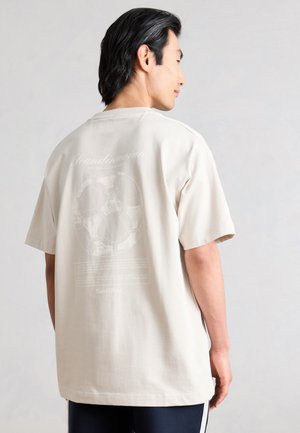 Άνδρας με μαύρα μαλλιά μεσαίου μήκους που στρέφεται πλαγίως, φορώντας χαλαρό μπεζ T-shirt με αχνό σχέδιο μπάλας ποδοσφαίρου στην πλάτη.