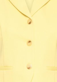 Blazer jaune avec un revers cranté, trois boutons beige et deux poches passepoilées à l'avant, présenté en gros plan.