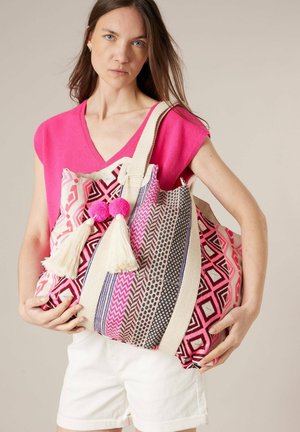 Femme portant un haut rose et un short blanc, tenant un grand sac fourre-tout à motifs avec des pampilles et des pompons.
