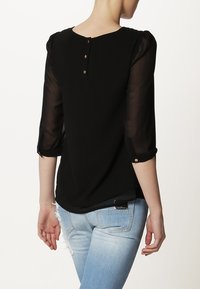 Svart blus med genomskinliga ärmar, rund halsringning och knapps detaljer på ryggen. Matchas med ljusblå slitna jeans. Slät textur.