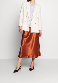 Blazer crema doppiopetto con bottoni dorati, top azzurro chiaro e gonna midi in raso color ruggine lucido, abbinati a décolleté nere con slingback.