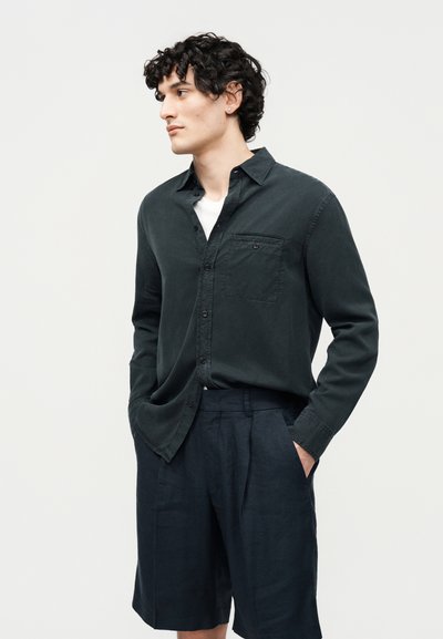 Homme aux cheveux bouclés portant une chemise boutonnée vert foncé sur un t-shirt blanc et un short foncé, debout les mains dans les poches, regardant vers la gauche.