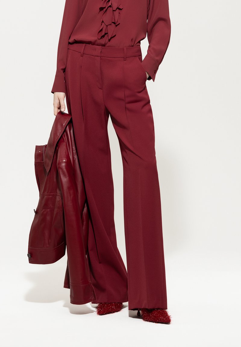 Burgunderfarbene, weit geschnittene Hosen mit Falten, kombiniert mit einer blusigen Bluse in passender Farbe und einer Lederjacke, alles in harmonierenden Farbtönen.