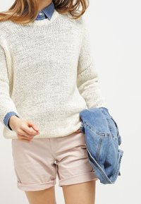 Světle krémový pletený svetr s kulatým výstřihem, přes modrou košilku, doplněný o růžové kraťasy. Modrá džínová bunda je přehozená přes rameno.