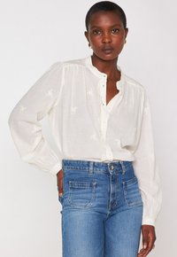 Donna con i capelli corti che indossa una camicetta bianca con bottoni e rouches e jeans a vita alta blu, in piedi con una mano sulla vita.