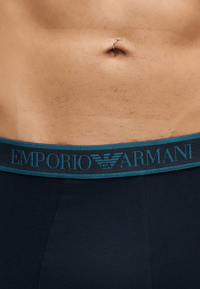 Gros plan d’un ventre nu avec un anneau de nombril en argent au-dessus de la ceinture d’un sous-vêtement sombre avec un logo Emporio Armani turquoise.