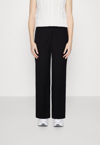 Une personne portant un pantalon noir à jambe large, un haut sans manches en tricot blanc et des chaussures de sport blanches, se tenant devant un fond uni.