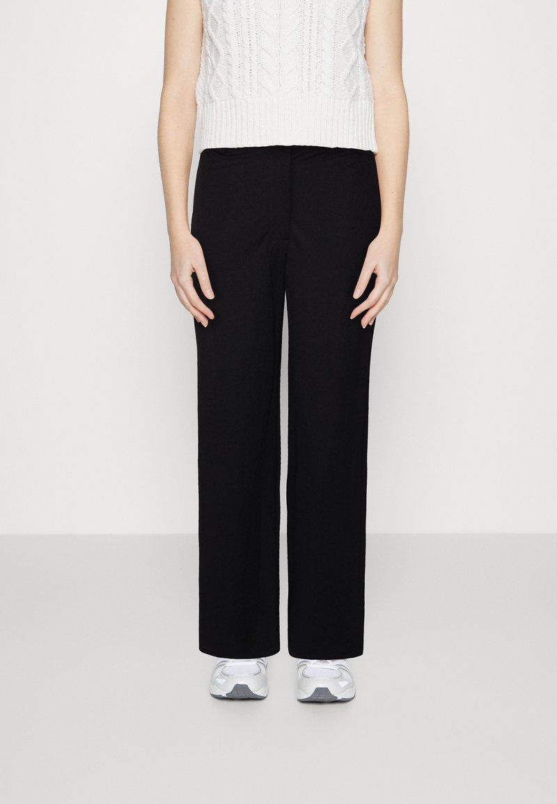 Une personne portant un pantalon noir à jambe large, un haut sans manches en tricot blanc et des chaussures de sport blanches, se tenant devant un fond uni.