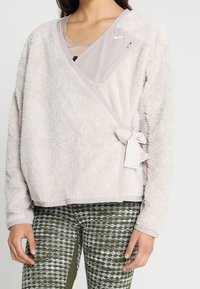 Kobieta ubrana w jasnobeżową polarową kurtkę z zamkiem błyskawicznym w kieszeni na piersi i związaną boczną kokardą, w połączeniu z zielonymi legginsami w wzory.