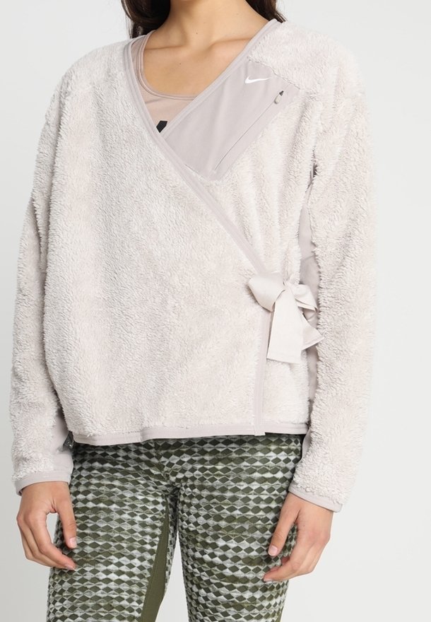 Kobieta ubrana w jasnobeżową polarową kurtkę z zamkiem błyskawicznym w kieszeni na piersi i związaną boczną kokardą, w połączeniu z zielonymi legginsami w wzory.