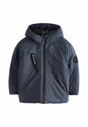 Junge mit lockigem blondem Haar, der eine schwarze, wattierte Winterjacke mit Kapuze und schwarze Hose trägt, steht und lächelt vor einem einfarbigen Hintergrund.