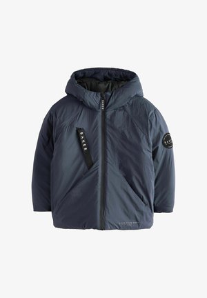 Junge mit lockigem blondem Haar, der eine schwarze, wattierte Winterjacke mit Kapuze und schwarze Hose trägt, steht und lächelt vor einem einfarbigen Hintergrund.