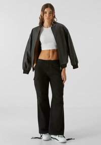 Svart läderbomberjacka, vit kortärmad top, svarta cargo-byxor med fickor, och vita sneakers med gröna accenter.