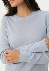 Világoskék kötött pulóver kerek nyakkivágással, bordás mandzsettákkal és sima textúrával, részben keresztbe tett karokat takar.