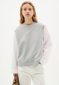 Felpa grigia in cotone con collo a giro e maniche lunghe in tessuto bianco, indossata sopra pantaloni chiari, abbinata a una piccola borsa tan.