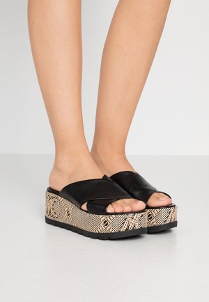 Feet wearing black leather crisscross strap platform sandals with woven patterned beige and black thick soles on a light floor.