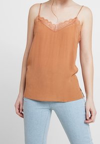 Rustrød cami-top med blondekant, justerbare spaghetti-stropper og en blød, glat tekstur. Bæres med lyseblå tætsiddende jeans.