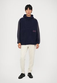 Sweater de capuz azul-marinho feito de tecido texturizado, com um bolso frontal, etiqueta roxa e riscas nas mangas, combinado com calças cremadas e sapatos pretos.
