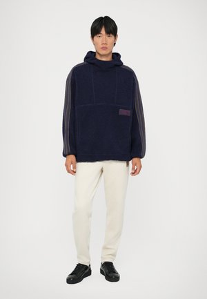 Sweater de capuz azul-marinho feito de tecido texturizado, com um bolso frontal, etiqueta roxa e riscas nas mangas, combinado com calças cremadas e sapatos pretos.