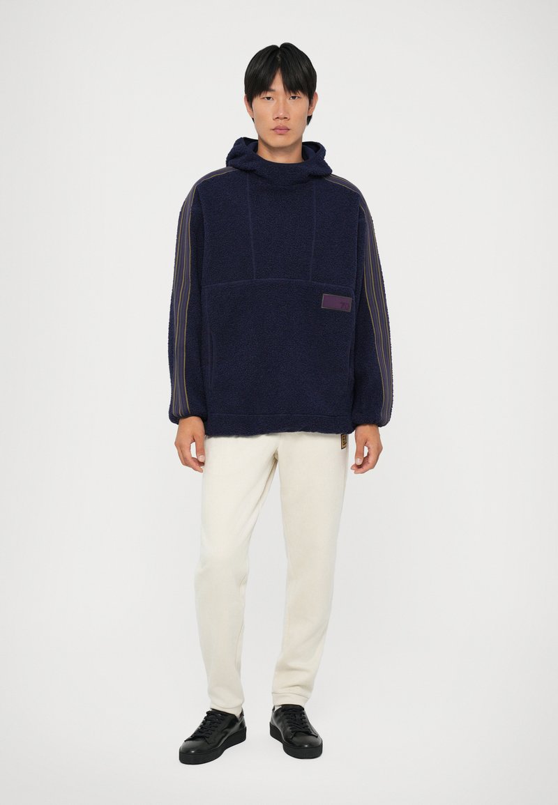 Sweater de capuz azul-marinho feito de tecido texturizado, com um bolso frontal, etiqueta roxa e riscas nas mangas, combinado com calças cremadas e sapatos pretos.