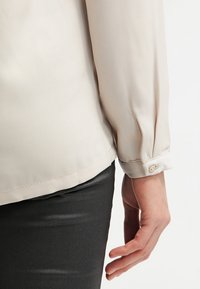 Blouse beige clair à manches longues avec des détails aux poignets et une texture de tissu douce et lisse. La chemise est assortie à un pantalon foncé et brillant.