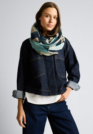 Młoda kobieta w niebiesko-beżowym wzorzystym szaliku, ciemnej dżinsowej kurtce z podwiniętymi rękawami oraz ciemnych dżinsach, stojąca na jednolitym tle.