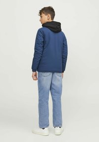 Blå vadderad jacka med svart huva, bärs �över ljusblå jeans och vita sneakers. Synlig sömnad och avslappnad passform.