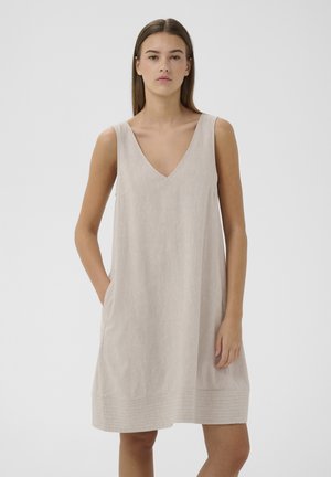 Joven mujer con un vestido sin mangas de lino beige con escote en V, de pie con una mano en el bolsillo contra un fondo blanco.