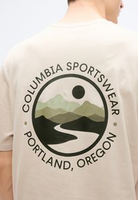 Μπεζ βαμβακερό μπλουζάκι με κυκλικό γραφικό που απεικονίζει βουνά, ένα ποτάμι και το κείμενο «Columbia Sportswear Portland, Oregon» σε σκουρό πράσινο χρώμα.