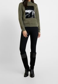 Sudadera verde oliva con estampado gráfico negro, combinada con jeans ajustados negros y botas negras de ante por encima de la rodilla con acabado brillante.