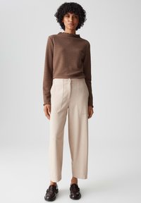 Bruine gestructureerde langε mouwtop met een gerimpelde halslijn, gecombineerd met beige wijde broek met voorzakken. Donkere schoenen maken de look compleet.