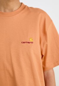 T-shirt en coton orange avec un col rond et des manches courtes. Le côté supérieur gauche de la poitrine présente un logo brodé jaune et rouge.