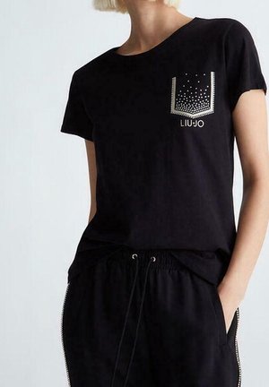 Kobieta ubrana w czarną koszulkę z krótkim rękawem z błyszczącym wzorem na kieszeni oraz dopasowane czarne spodnie z wiązanym paskiem w pasie i zamkami po bokach.