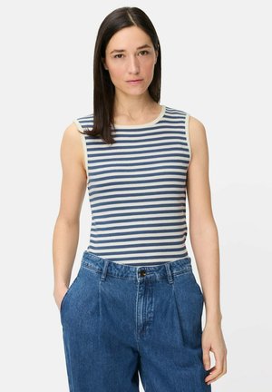 Kvinde med mørkt hår iført en ærmeløs blå- og hvidstribet top, stoppet ned i højtaljede, løstsiddende blå jeans, hænder i lommerne, neutral baggrund.