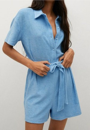 Woman wearing a light blue short-sleeve button-up romper with a collar, tying a fabric belt around the waist.