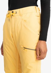 Gelbe Hose aus leichtem Material, mit einem Kordelzugbund, schrägen Taschen und einer Reißverschlusstasche an der Seite.