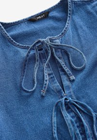 Haut en denim avec un col rond, un détail à nouer à l'avant, et un tissu bleu avec des coutures visibles. La texture est lisse et légèrement délavée.