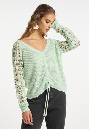 Frau trägt einen hellgrünen gestrickten Pullover mit Häkelärmeln und einem vorderen Kordelzug, kombiniert mit dunklen, hoch taillierten Hosen.