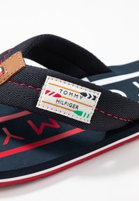 Dunkelblaue Flip-Flops mit strukturierten Stoffriemen, roter Naht und braunem Lederakzent. Verfügt über ein Etikett mit „TOMMY HILFIGER“-Markenaufdruck.