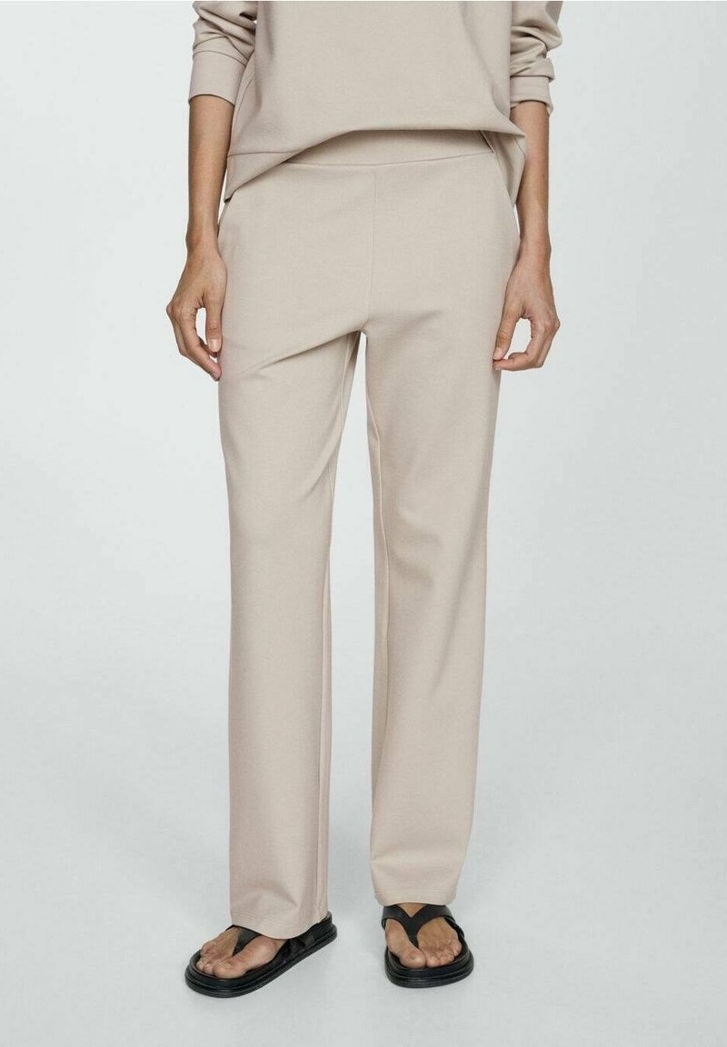 Personne portant un pantalon beige à jambes droites et des sandales noires à brides, debout devant un fond clair uni.