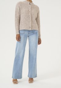 Mujer vistiendo un cardigán de punto beige abotonado, jeans de pierna ancha de color azul claro y sandalias abiertas de dedo beige, de pie sobre un fondo blanco.