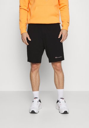 Mężczyzna ubrany w czarne szorty Champion, białe buty Nike, białe skarpetki oraz pomarańczową bluzę, stoi na tle jednolitego jasnego tła.