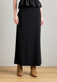 Jupe noire, longueur genoux, en tissu lisse ; design ajusté avec un ourlet évasé ; associée à des bottines en daim tan à la cheville.